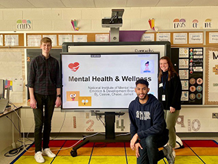 Three individuals in an elementary school classroom surround a presentation board. The board has heart, meditation, and facial expression emojis and reads Mental Health & Wellness, National Institute of Mental Health, Emotion & Development Branch, Bj, Cassie, Chase, Jamell. 