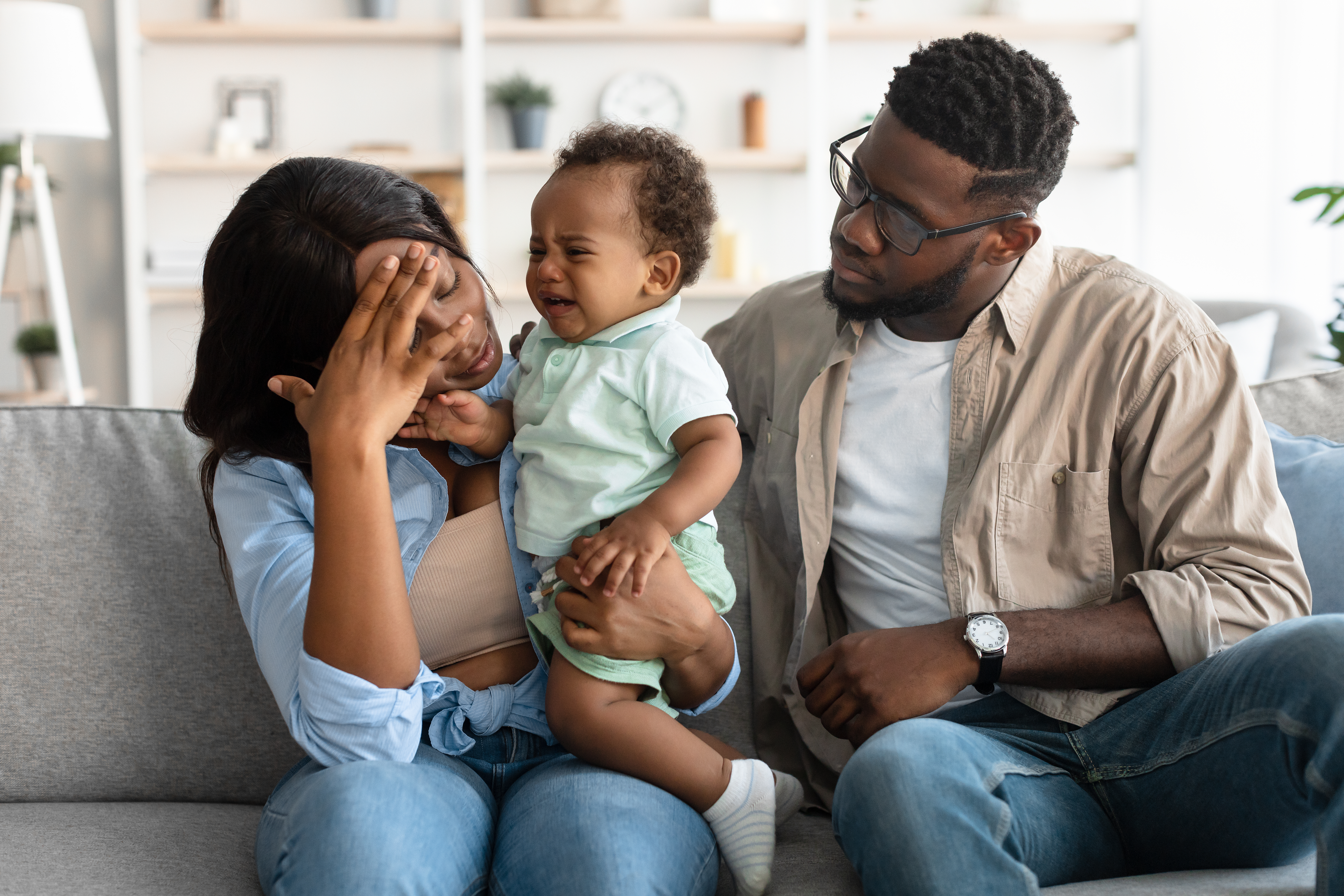 Mom with head in hand sitting on couch and holding crying baby, while dad sits beside them and looks on with worry.. 