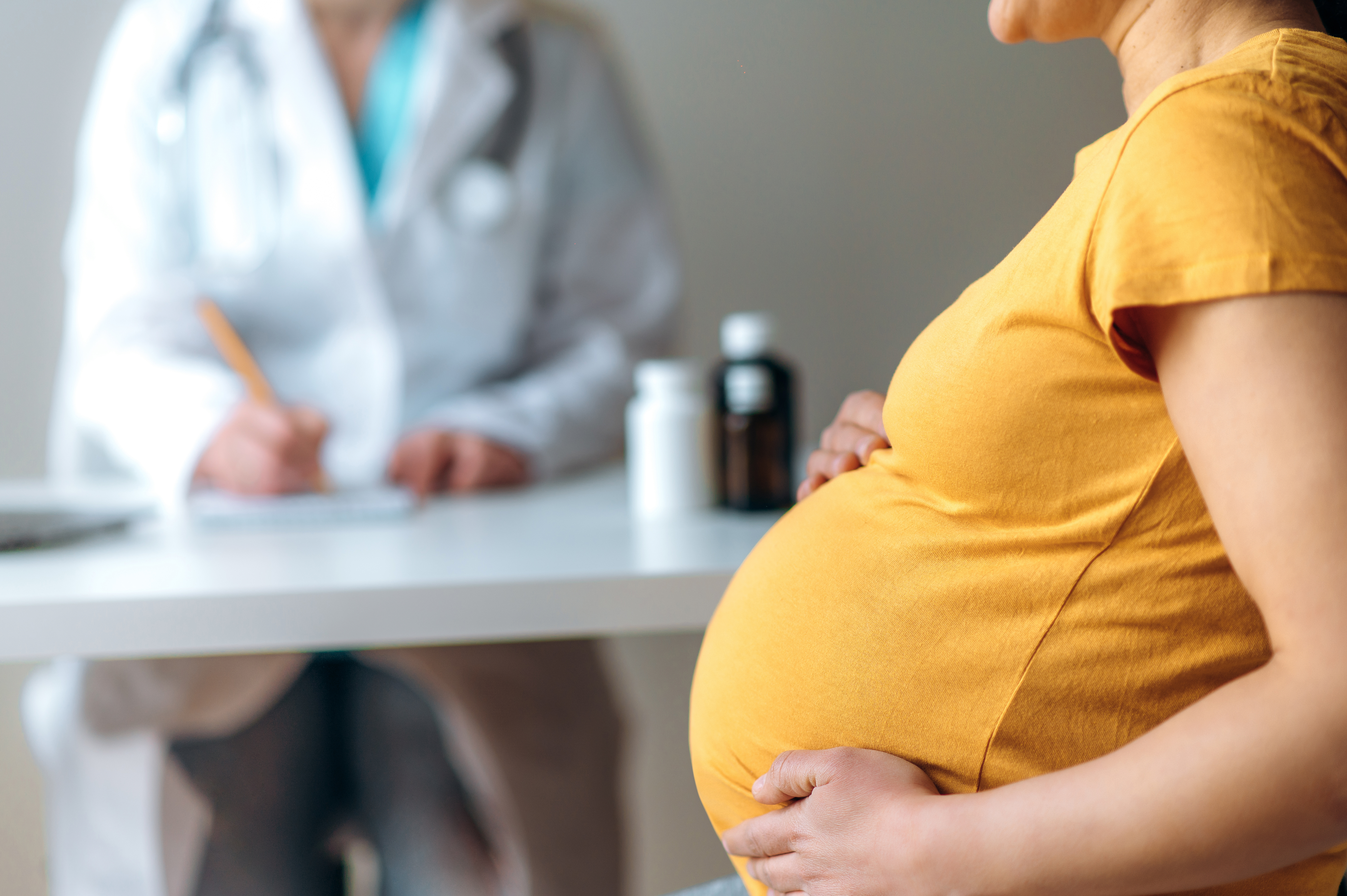 Profile shot of woman holding her pregnant belly with a doctor writing a prescription in the background.