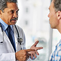 A doctor wearing a white lab coat and stethoscope facing a male patient wearing a white and blue flannel shirt.