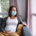 Worried pregnant woman sitting at home with protective face mask, looking through the window and holding stomach.