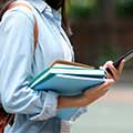 A student carries books and a phone on a college campus. 