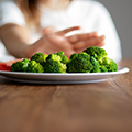 A teen (in background) pushes away a plate with broccoli on it (in foreground).
