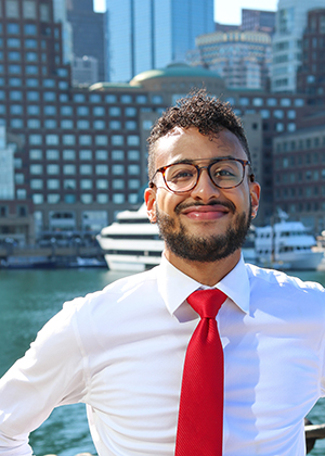 Dressed in a white shirt and red tie, Josh Santana smiles with the Boston cityscape behind him. 