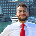 Dressed in a white shirt and red tie, Josh Santana smiles with the Boston cityscape behind him. 