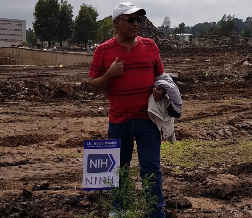 Abera Wouhib, Ph.D. stands in an open field , behind a small sign with the NIH logo and NIMH written on it.