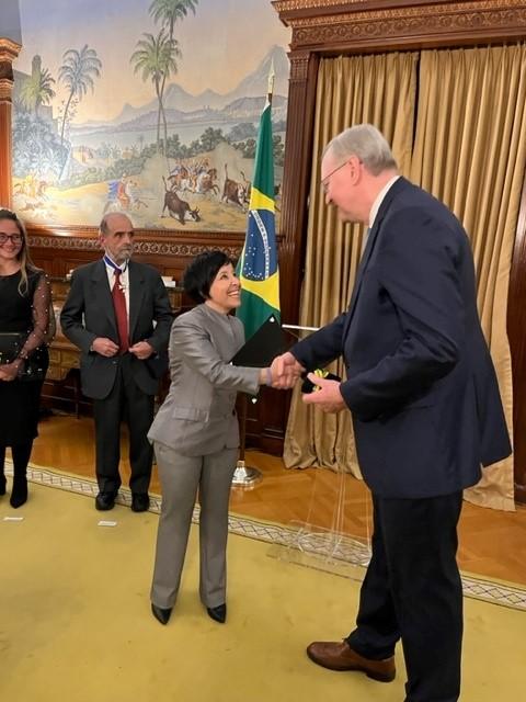 A woman in a gray suit, Andrea Horvath Marques, M.D., Ph.D., MPH, shakes hands with Ambassador Forster while receiving the Science and Technology Prize of the Embassy of Brazil.