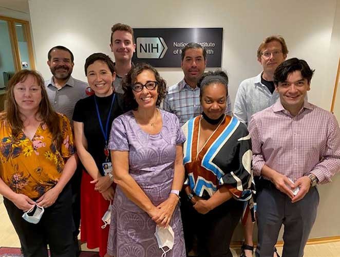 Devora Kestel, MSc, wearing a purple dress, stands in the middle of a group of 8 NIMH Global Mental Health team members. 